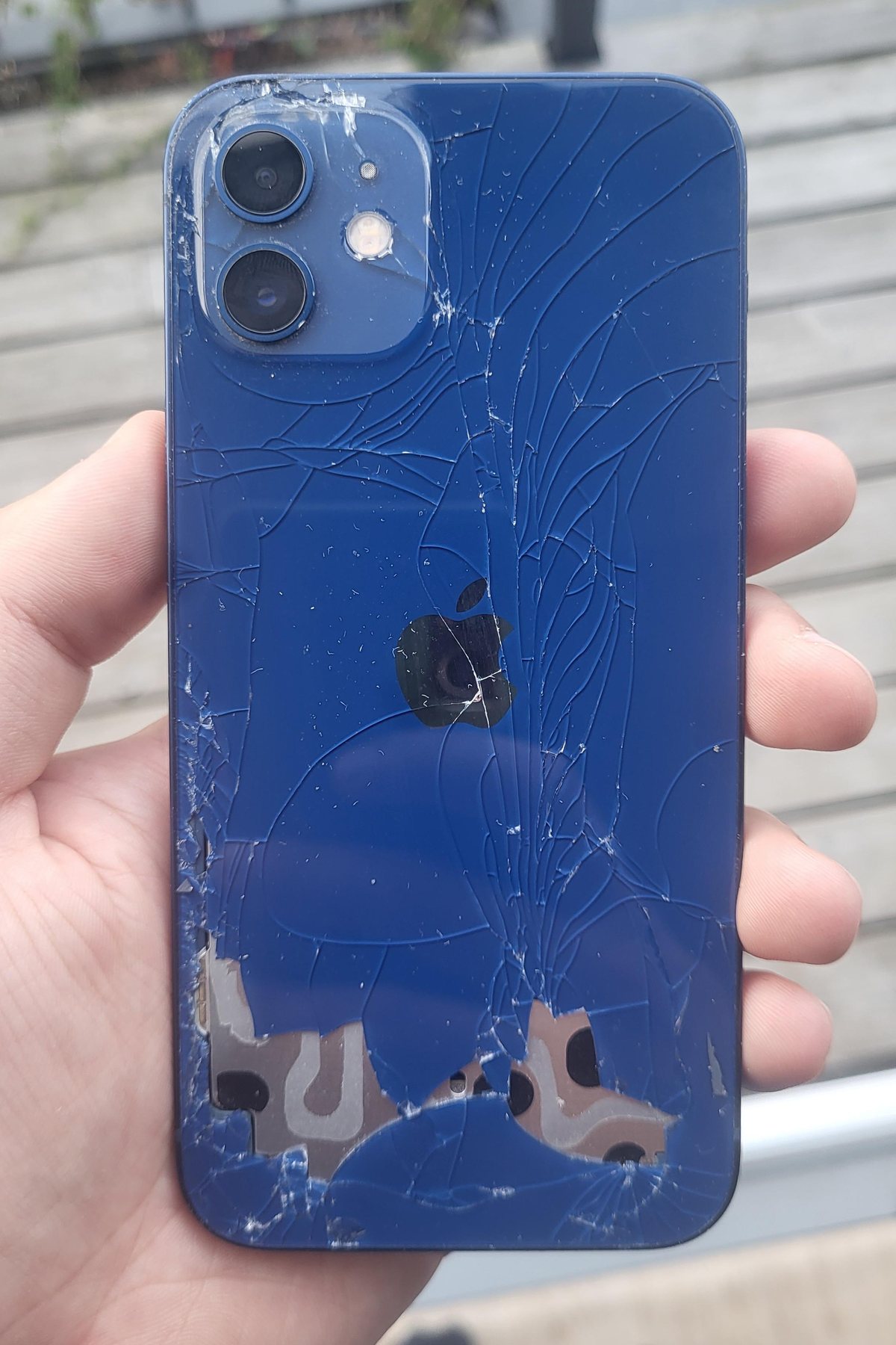 A blue iPhone held in a hand. The back glass is shattered and pieces are missing.